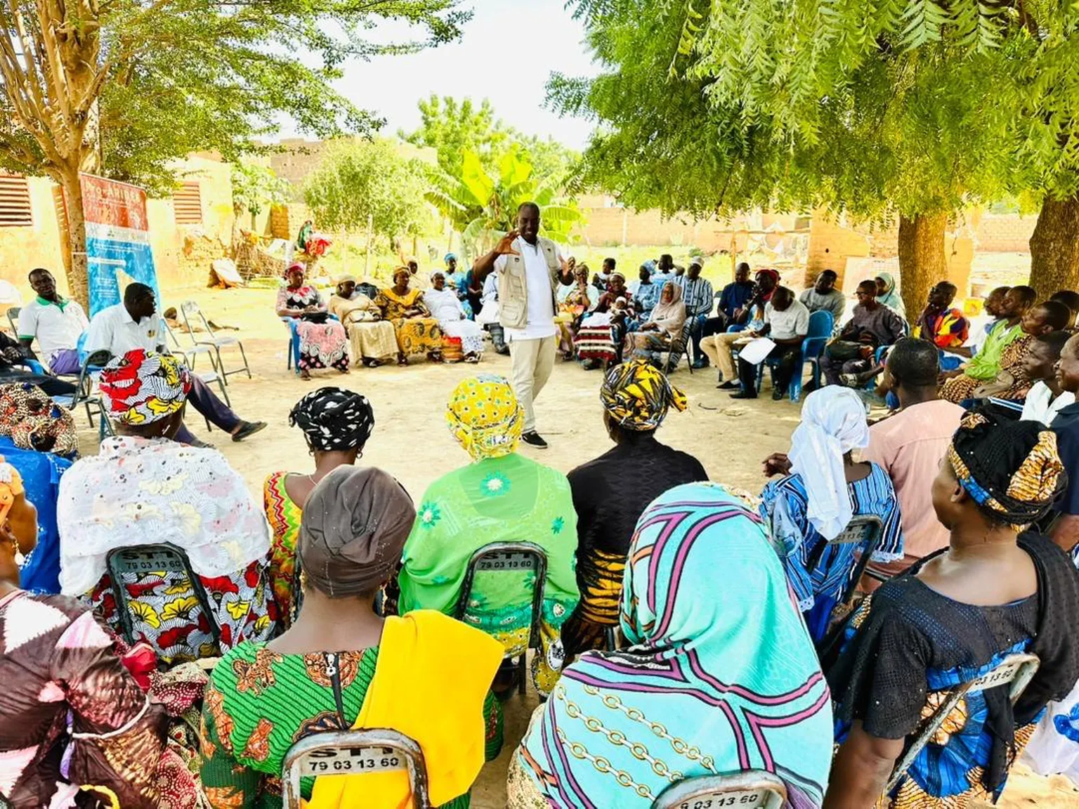 A diverse group of people sit in a circular outdoor meeting, engaged in discussion under the shade of large trees.