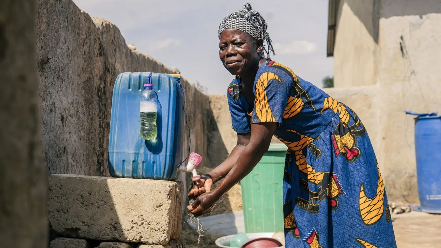 Woman washing hands at an outdoor faucet beside large water containers, wearing a blue patterned dress and headscarf, under a sunny sky.