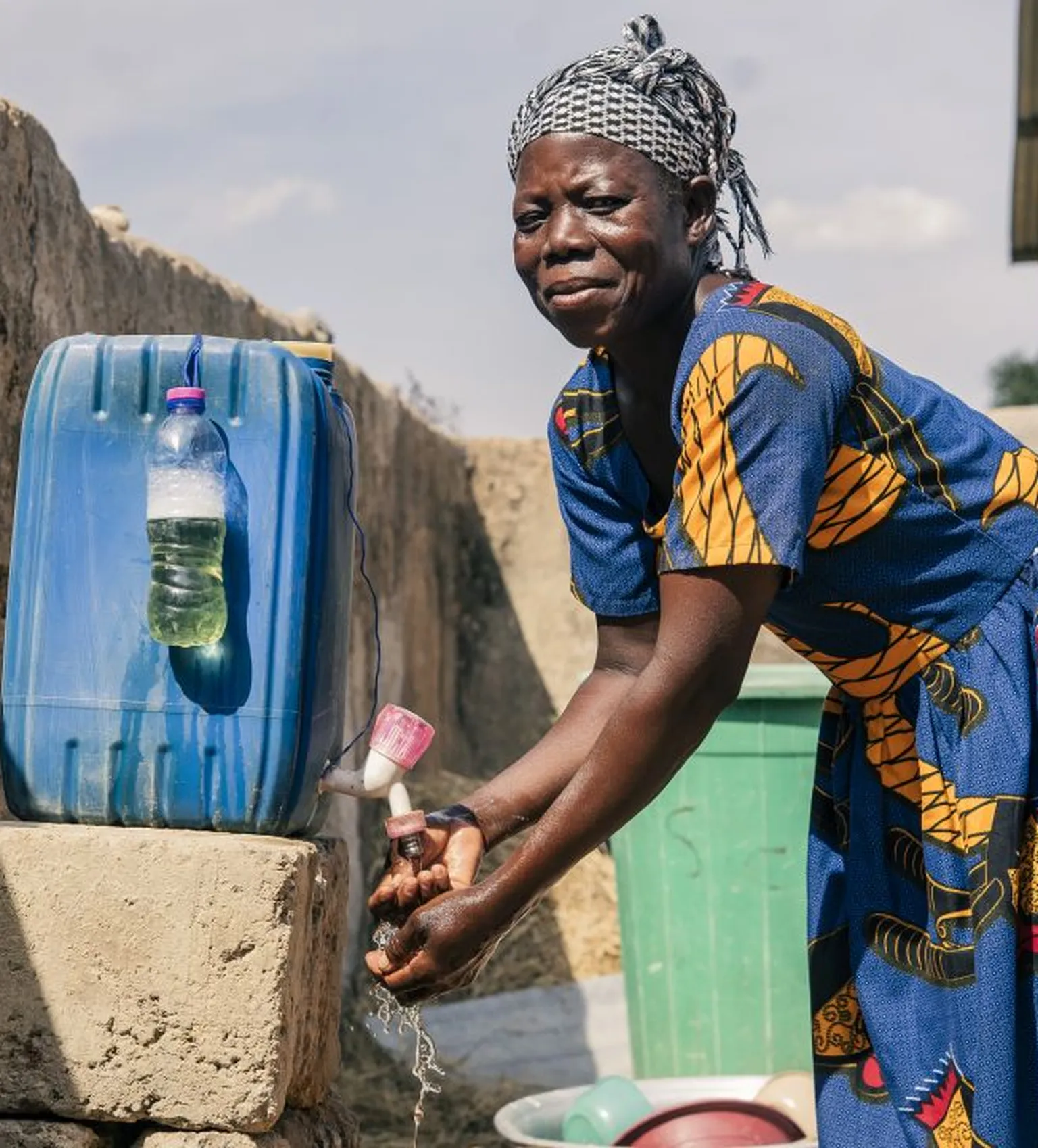 Woman washing hands at an outdoor faucet beside large water containers, wearing a blue patterned dress and headscarf, under a sunny sky.