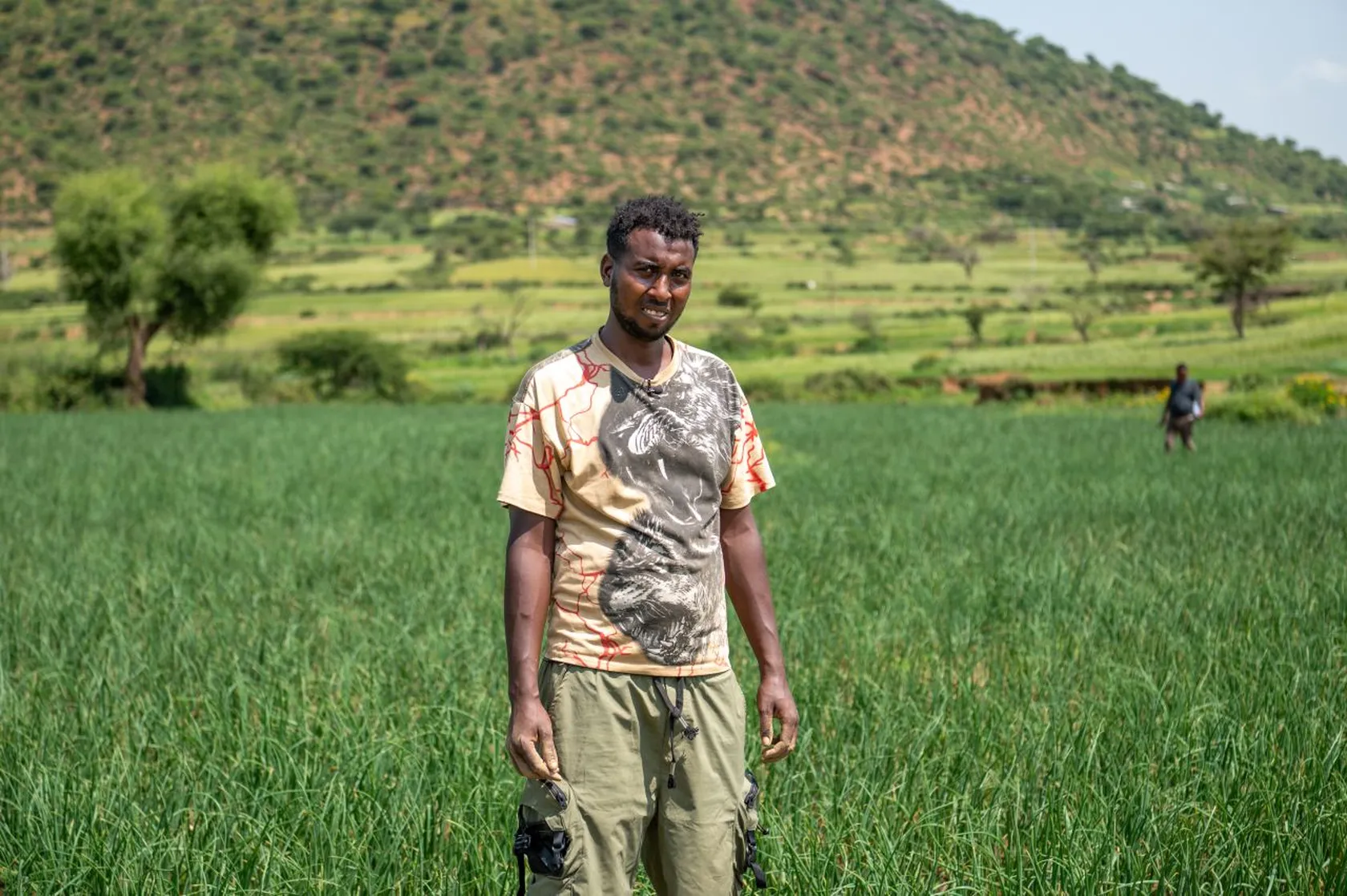A man stands in a lush green field, with a mountainous landscape in the background under a clear sky.