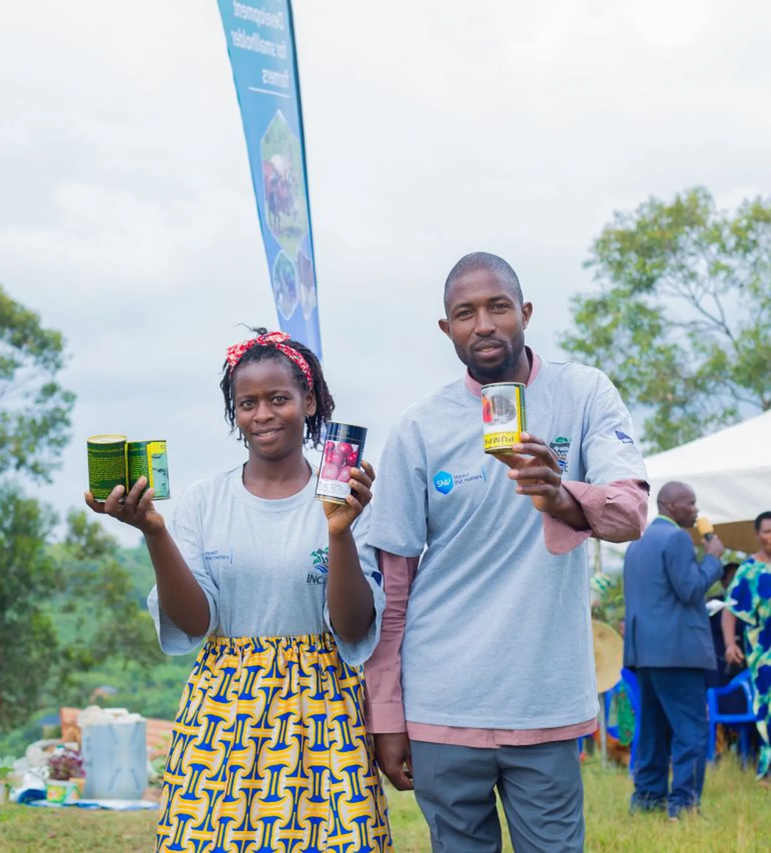 Two people stand outdoors, smiling and holding packaged products. A tent and several people are visible in the background.