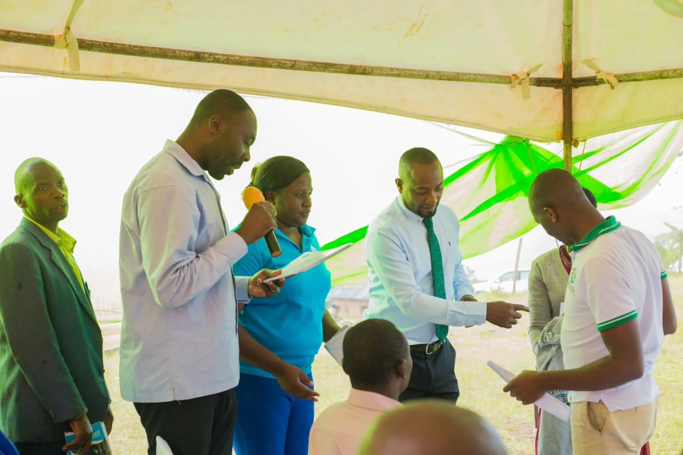 Group of people standing under a tent, some holding papers and a microphone, engaging in a discussion or presentation.