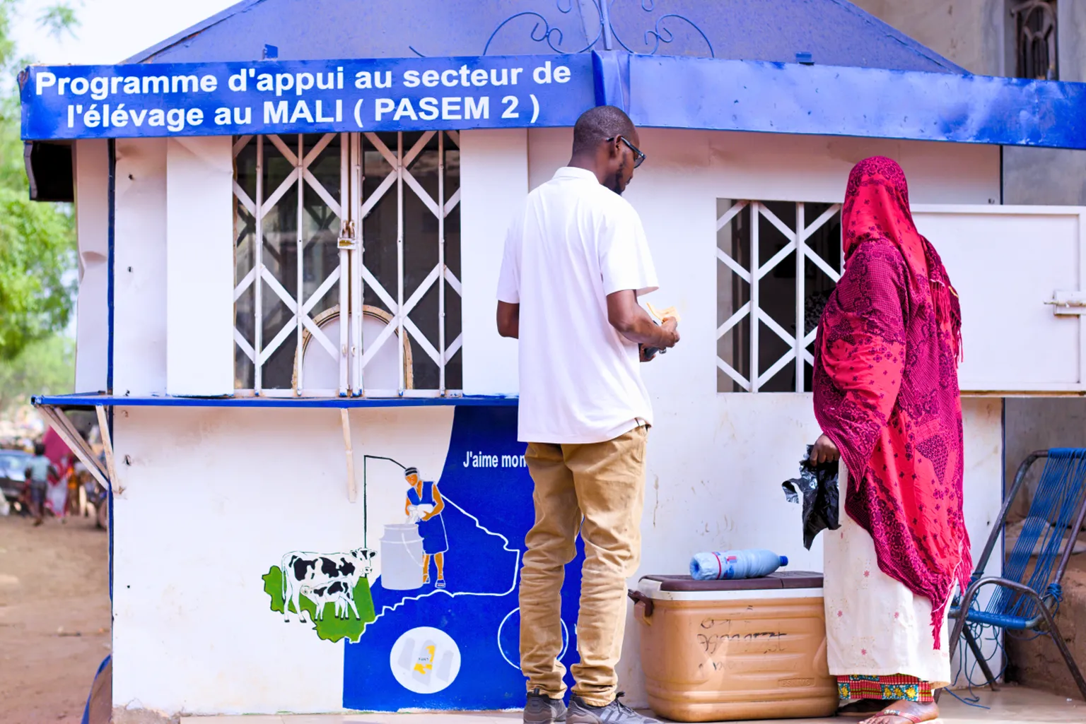 Two people stand outside a kiosk promoting Mali's livestock support program, with a colorful mural and blue signage above.