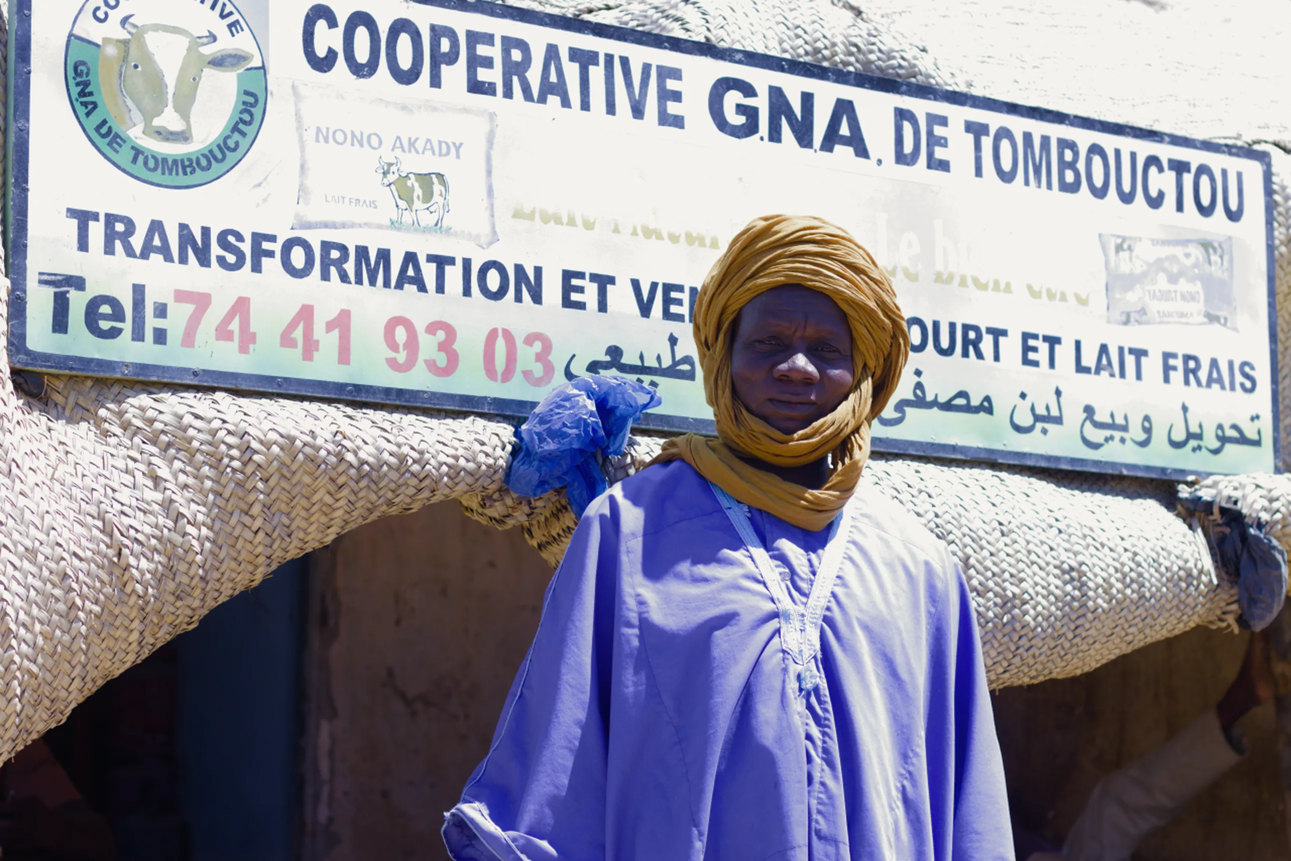 A person in traditional attire stands in front of a sign for Cooperative GNA de Tombouctou, related to milk processing and selling.