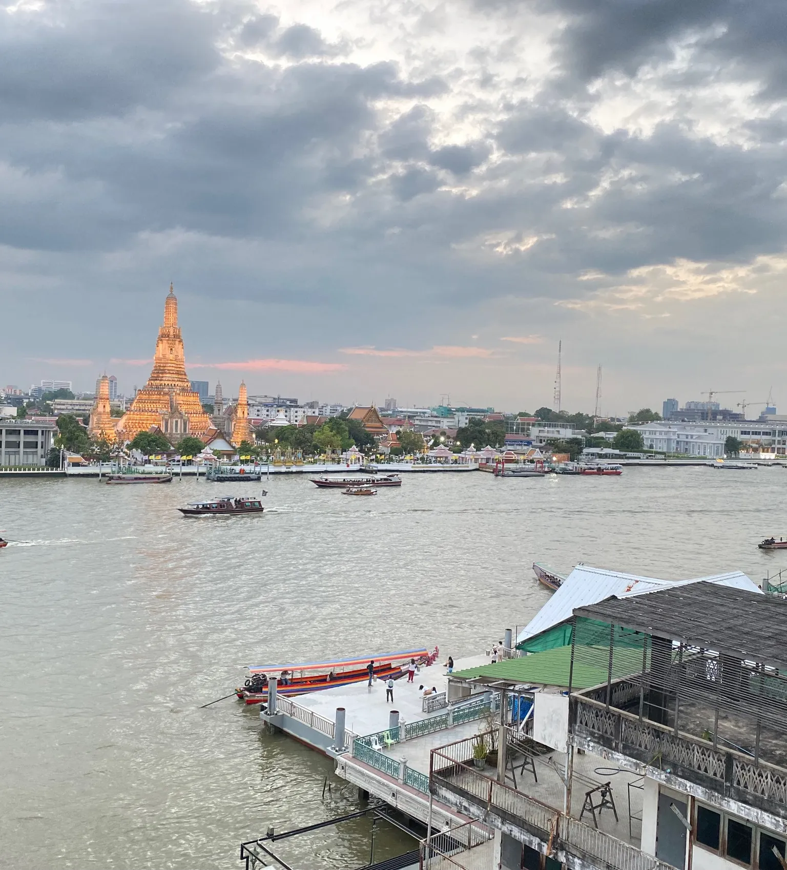 View of a river with boats, a golden temple on the opposite bank, and a cloudy sky at sunset.