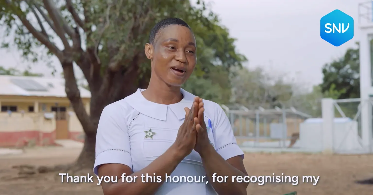 A smiling nurse in a white uniform, standing outdoors, clasps her hands while expressing gratitude. The SNV logo is visible at the top.