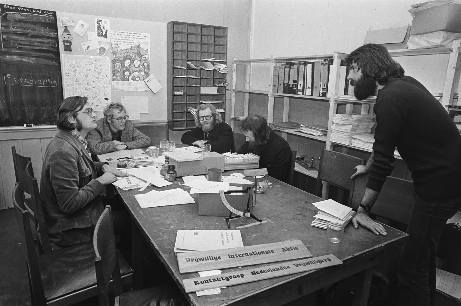 Five people seated around a cluttered table in an office, engaged in discussion. Shelves with files and posters are visible in the background.