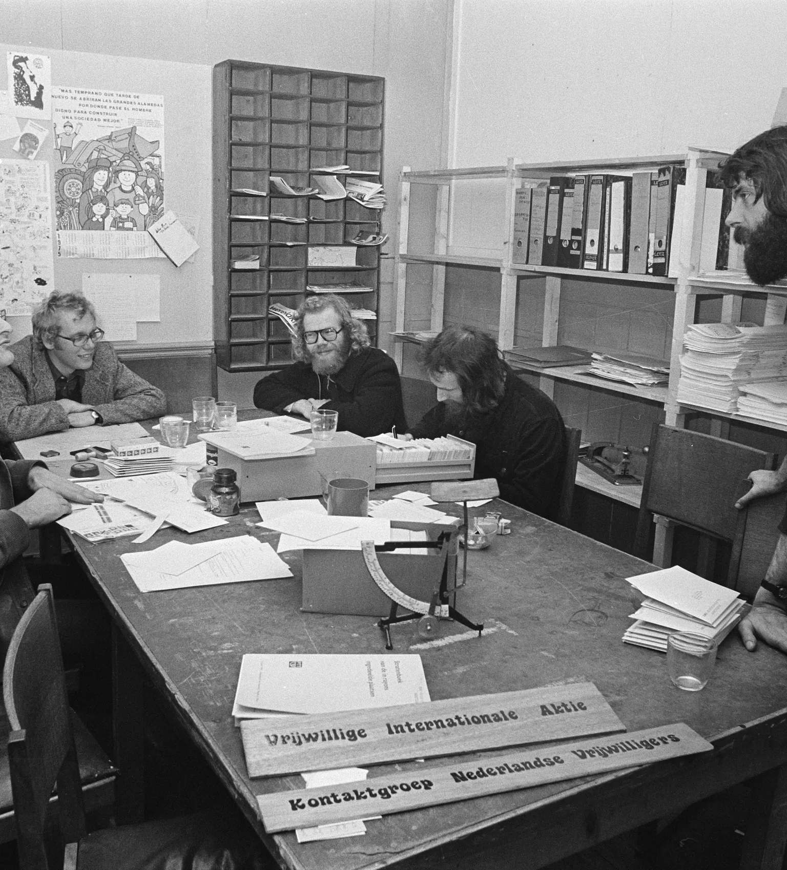 Five people seated around a cluttered table in an office, engaged in discussion. Shelves with files and posters are visible in the background.