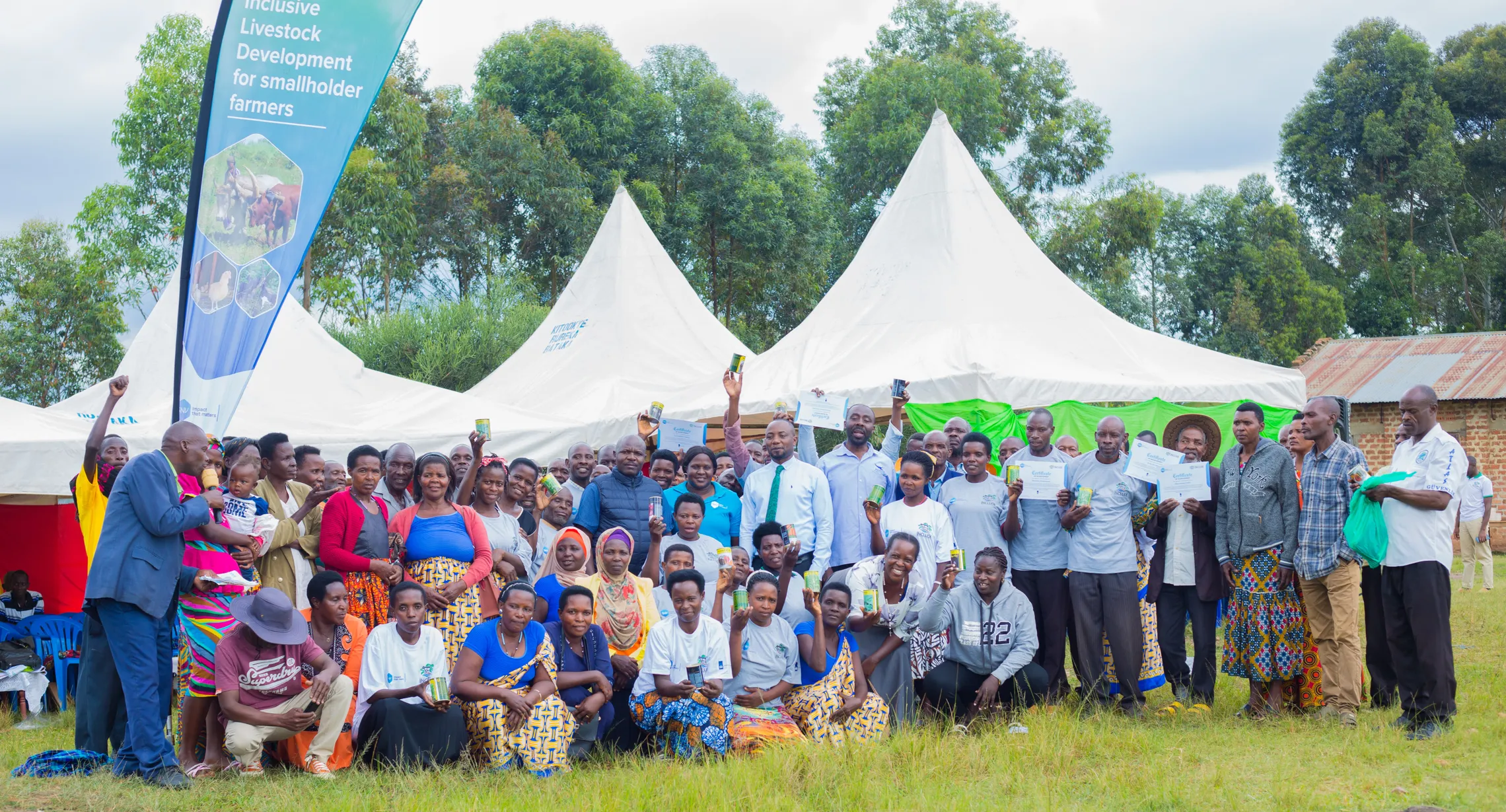 A large group of people pose outdoors in front of tents, holding certificates, with a banner about inclusive livestock development.