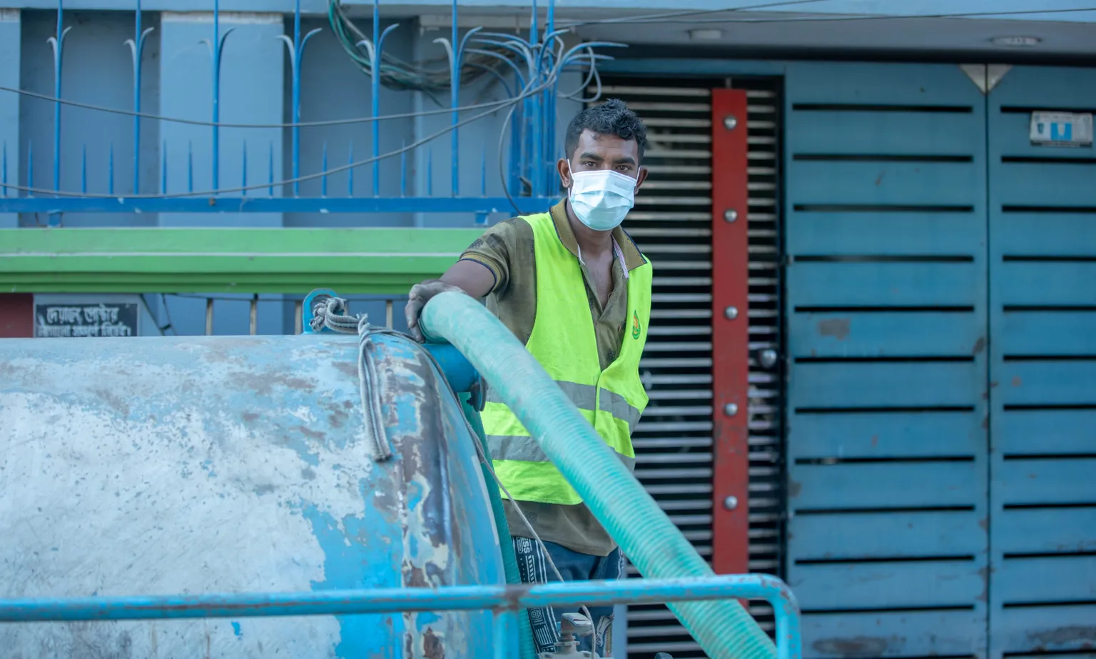 A worker wearing a mask and neon safety vest operates machinery with a large hose, standing in front of a blue and metallic striped garage door.