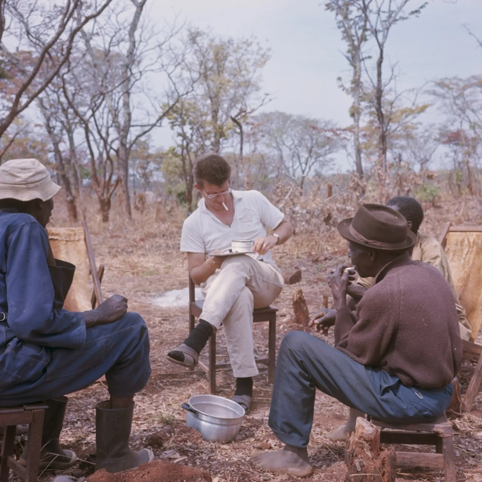 A person in a white shirt writes on a notepad, sitting with three others in a forested area, surrounded by dry grass and trees.
