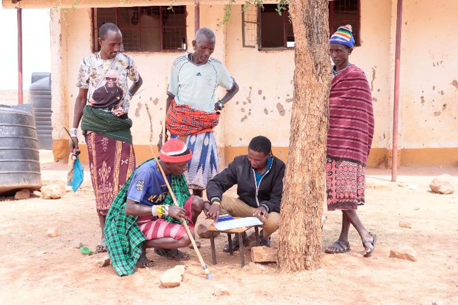 Five men in traditional clothing gathered under a tree by a building; one seated signs papers on a table while others watch
