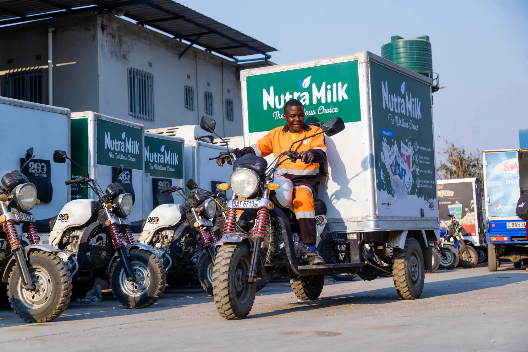 Man in orange uniform riding a NutraMilk delivery tricycle, surrounded by similar vehicles in a lot.