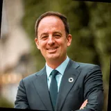 Smiling man in a suit and tie stands outdoors with arms crossed, wearing a circular pin on his lapel. Background is blurred greenery.