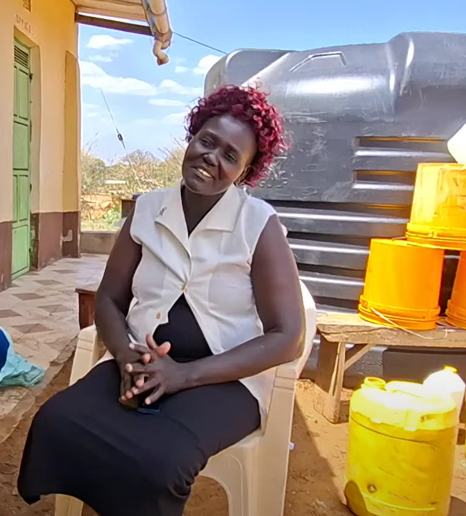Woman sitting outdoors on a plastic chair, smiling. Behind her are a large water tank and several orange and yellow jerry cans.
